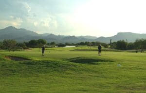 Two people standing on a green with mountains in the background.